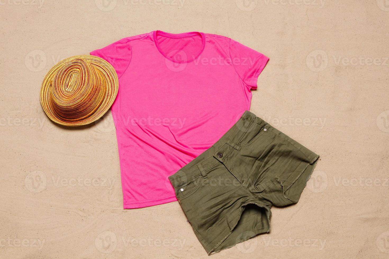 Sand beach texture background. Mockup pink summer t-shirt outfit copy space. Blank template woman shirt Top view. Summertime accessories hat, jeans. Flat-lay closeup tshirt on seashore. Beachtime photo