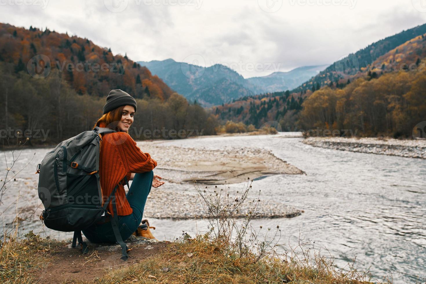 woman in the mountains in autumn with a backpack Sits on the river bank and looks at the high mountains photo
