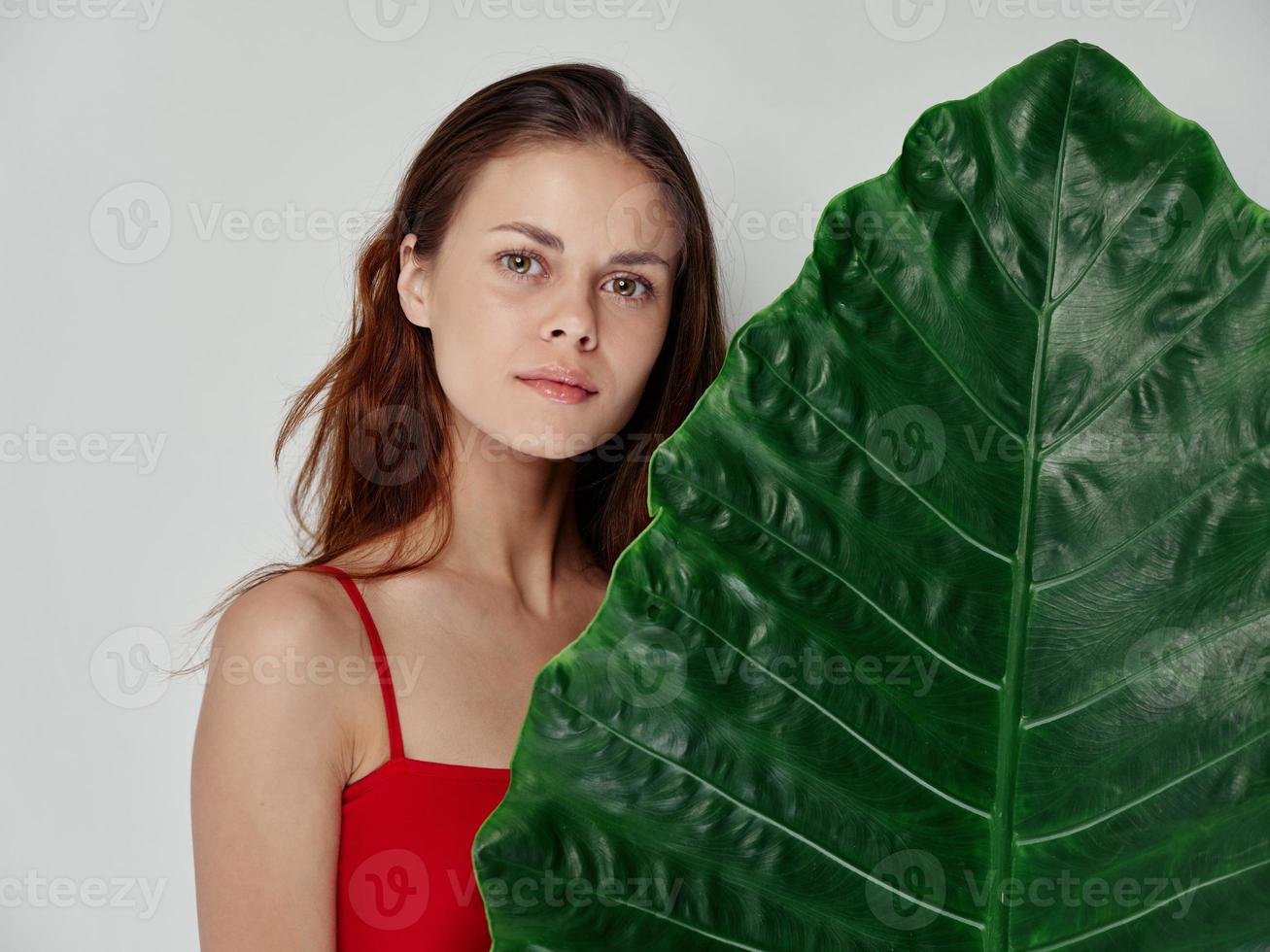 beautiful woman in a red t-shirt stands behind a green leaf of a palm tree on a light background clean skin photo