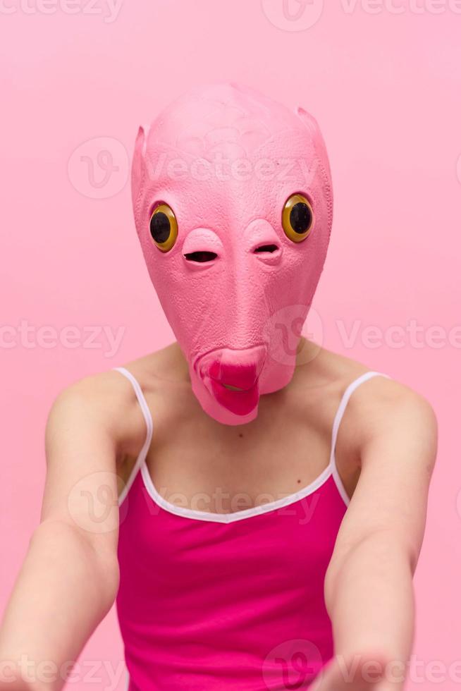 A woman wearing a silicone Halloween mask in the shape of a pink fish with big yellow eyes looks at the camera against a pink background photo