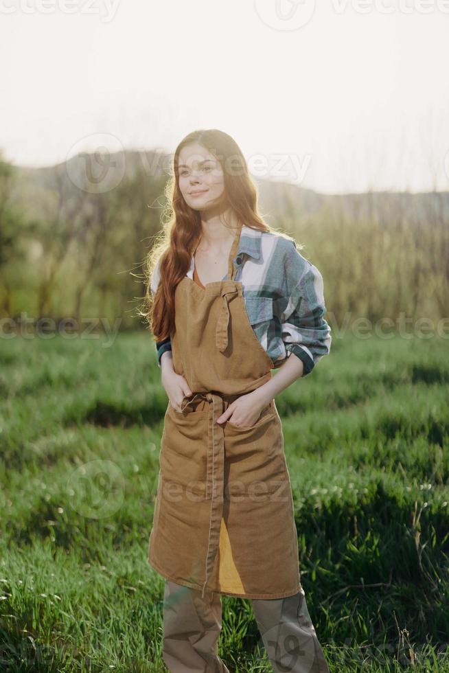 A farmer woman in her work clothes, plaid shirt and apron, stands in the field on the green grass and smiles in the setting sun after a hard day's work photo