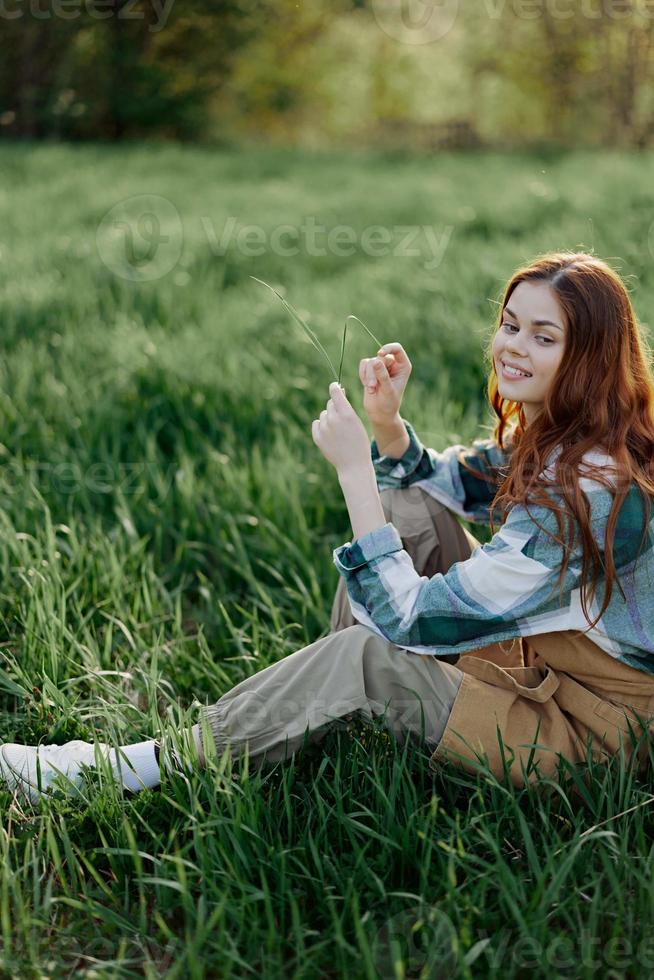 A woman sits on the green grass in a park with her back to the camera and relaxes in nature in the summer sunset evening light photo