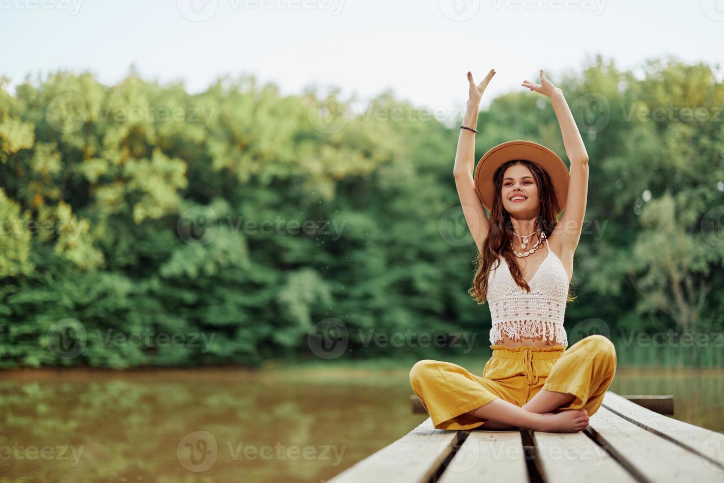 Hippie eco-activist woman traveler sits on a bridge by a lake with her arms outstretched with a hat and smiling sincerely photo