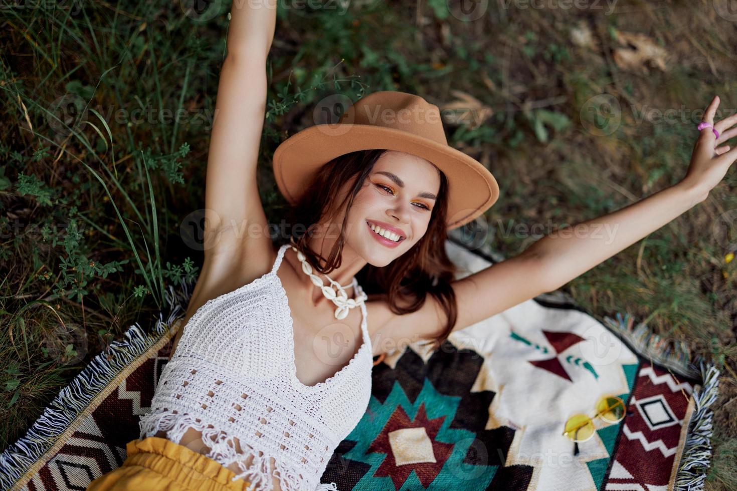 A woman in eco-friendly clothing in a hippie look is lying on a colored plaid smiling and looking at an autumn sunset in nature in the park. Lifestyle on a journey of harmony with the world photo