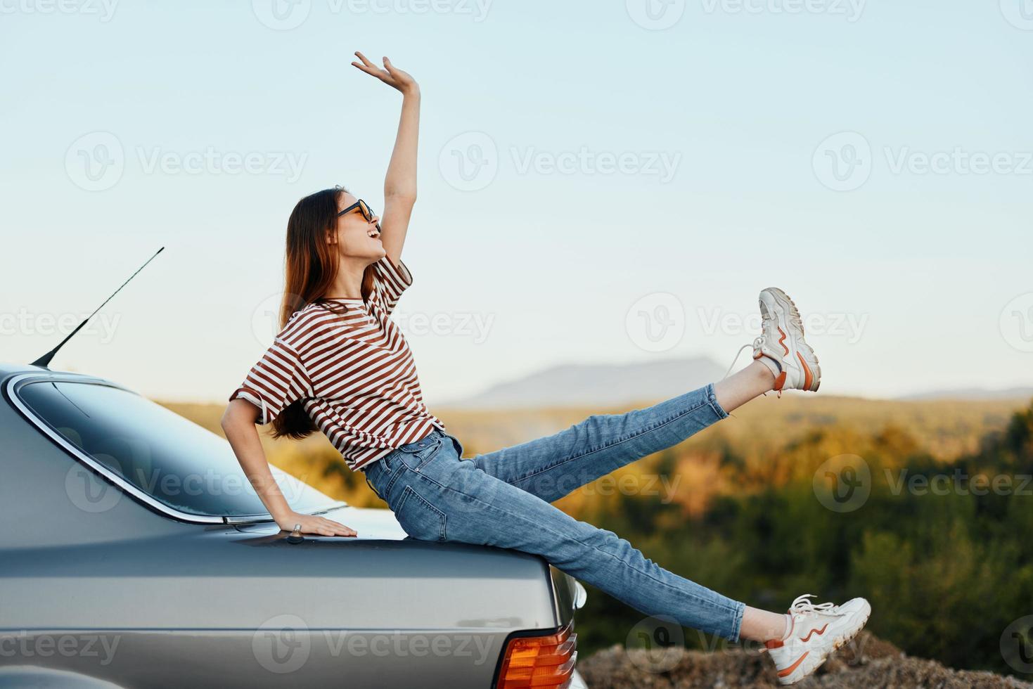 un mujer con un coche detenido en el la carretera a descanso en el viaje elevado su brazos y piernas desde felicidad y un hermosa paisaje foto