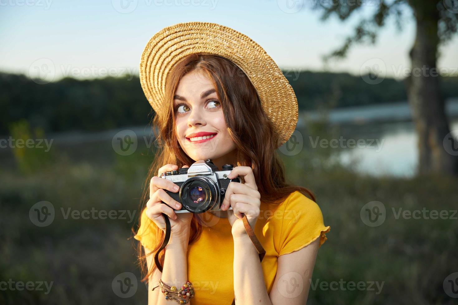 A woman photographer with a camera in her hands looks to the side bites her lip in a hat on the background of nature photo