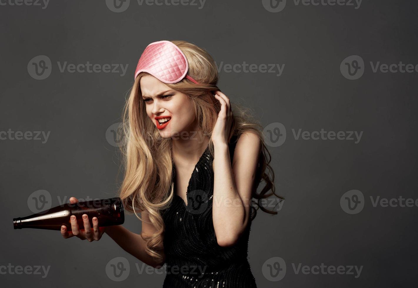 Drunk woman with a bottle of alcohol in her hand on a gray background and a mask for sleeping photo