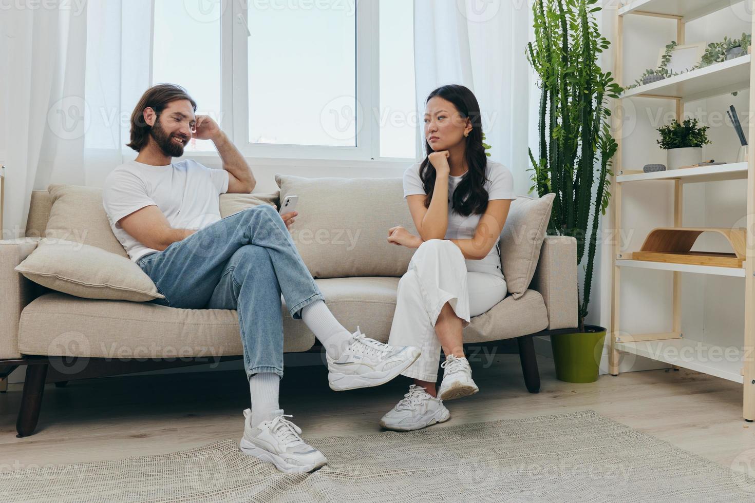 A man looks at his phone screen and smiles during an argument with his girlfriend. The angry and hurt woman looks in his direction and is sad. Family discord at home, phone addiction photo