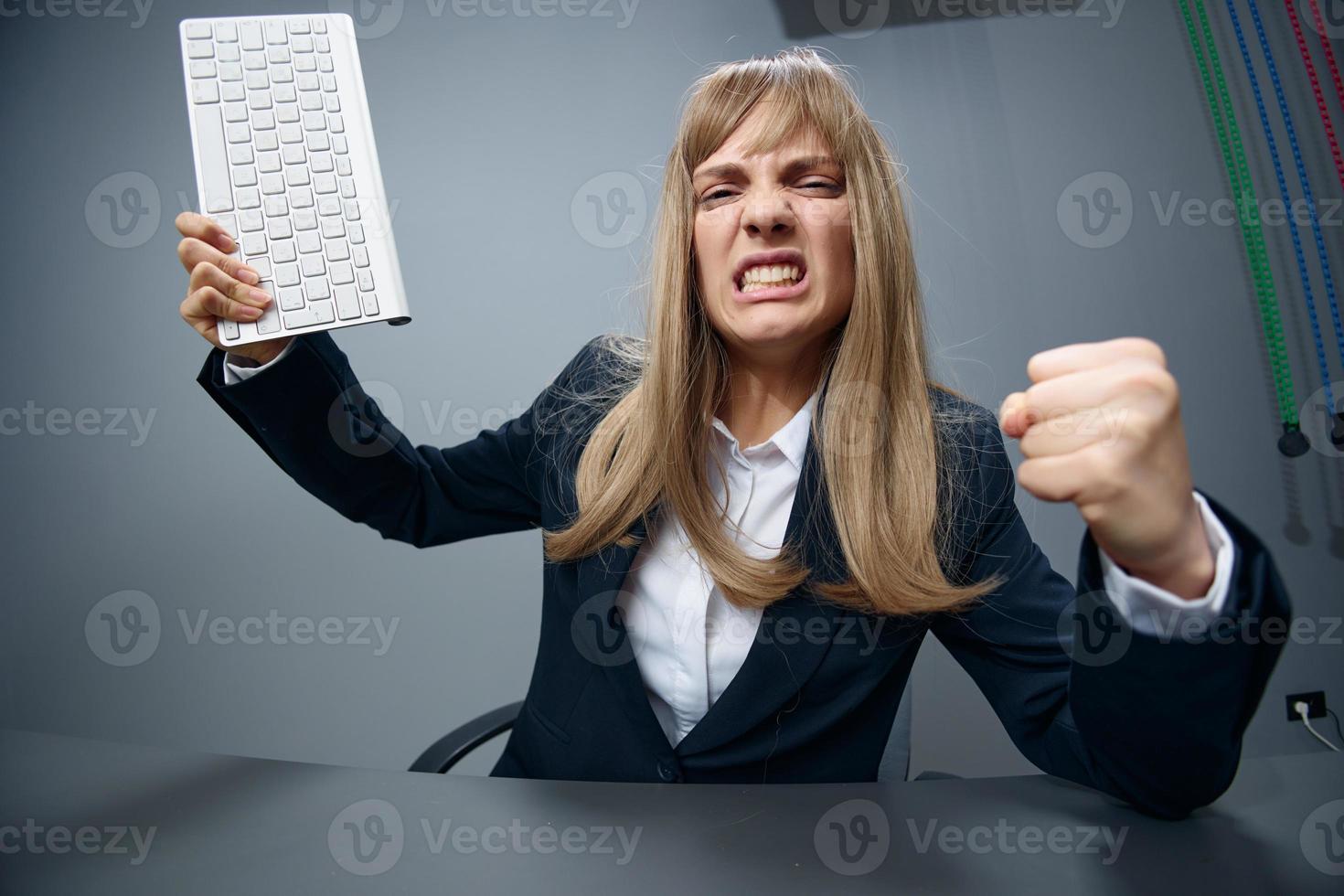 Crazy angry millennial blonde businesswoman worker in blue jacket throws keyboard and threatens at camera with fist in gray modern office. People Emotions Business Concept. Copy space, wide angle photo