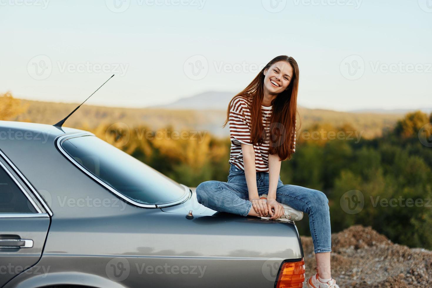A woman car driver sits on the trunk of a car and smiles admiring a beautiful view of autumn nature and mountains photo
