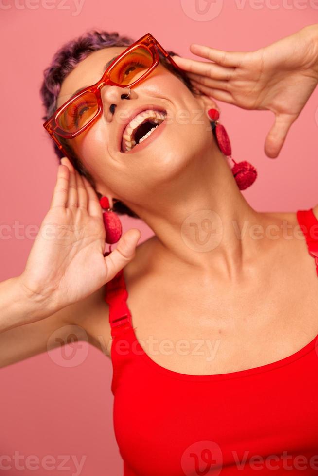 Young athletic woman with a short haircut and purple hair in a red top and pink yoga leggings in sunglasses with an athletic figure smiles and dances on a pink background photo