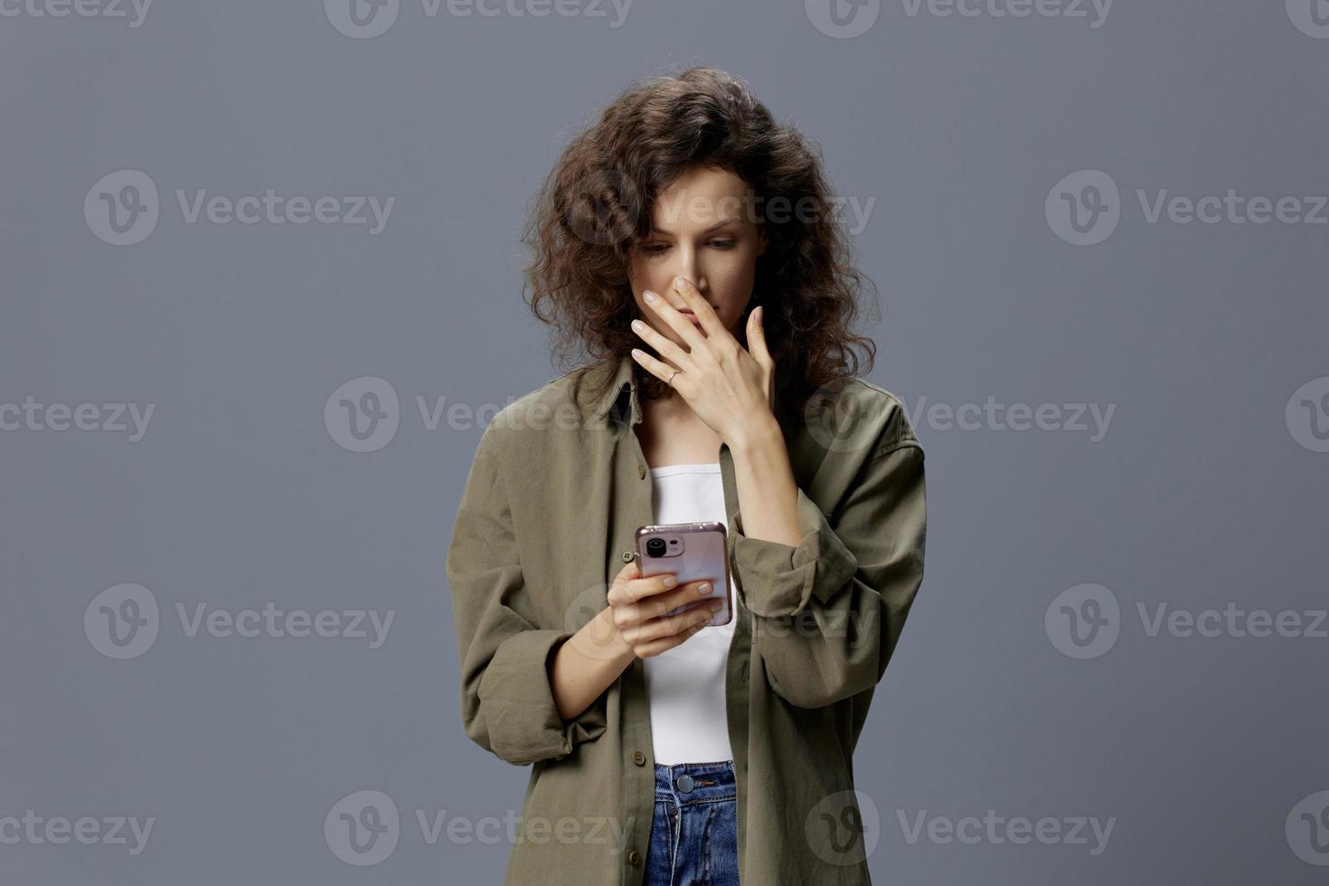 Upset shocked confused curly beautiful woman in casual khaki green shirt receiving bad news using phone posing isolated on over gray blue background. Social Media Influencer concept. Copy space photo