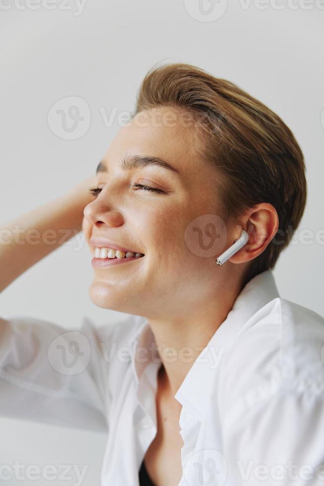 Young woman teenager listening to music with infertile headphones and dancing home, grinning with teeth with a short haircut in a white shirt on a white background. Girl natural poses with no filters photo