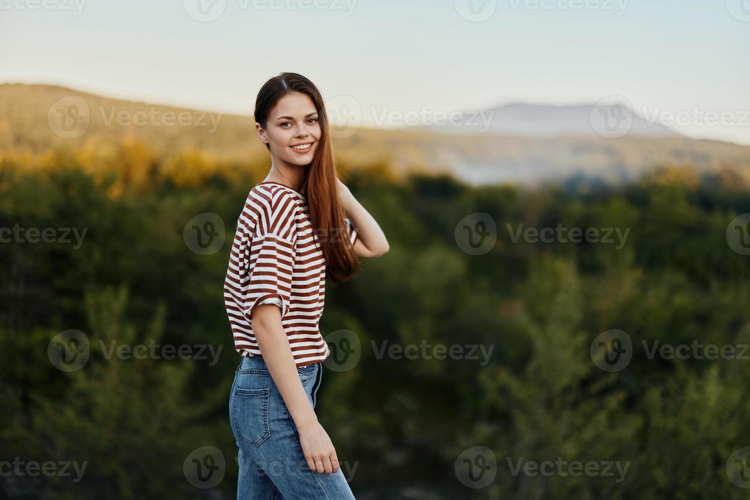 A young woman laughs and looks at the camera in simple clothes against the backdrop of a beautiful landscape of mountains and trees in autumn. Lifestyle on the move photo