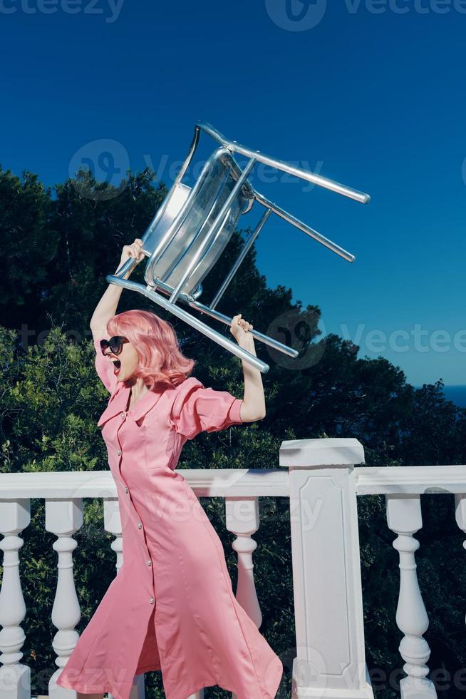 woman with pink hair in glasses with a chair in her hands on the balcony photo