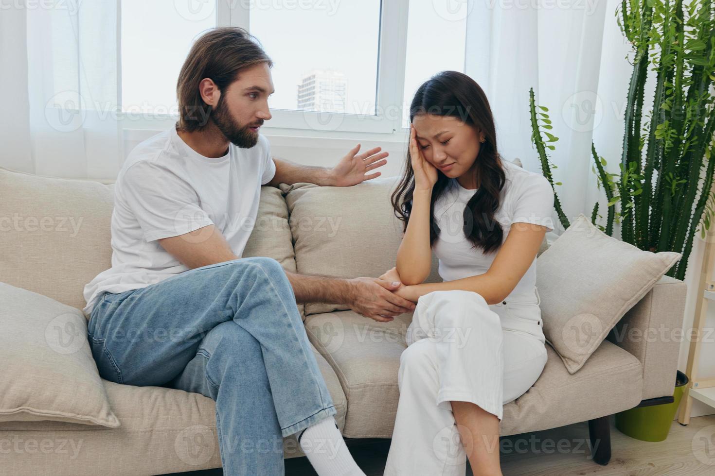 A sad Asian woman talks to a man in tears at home sitting on the couch. Young couple of different nationalities and conflict of interest in a couple photo