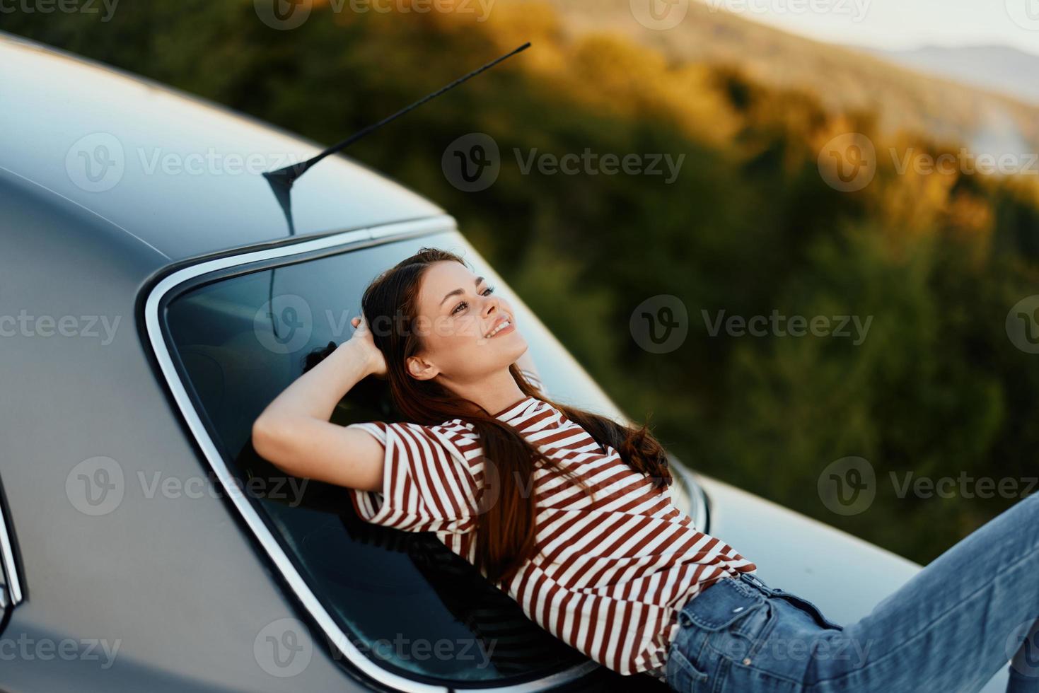 The woman driver stopped on the road and lay down on the car to rest and look at the beautiful landscape in a striped T-shirt and jeans. Complicated journey to nature photo