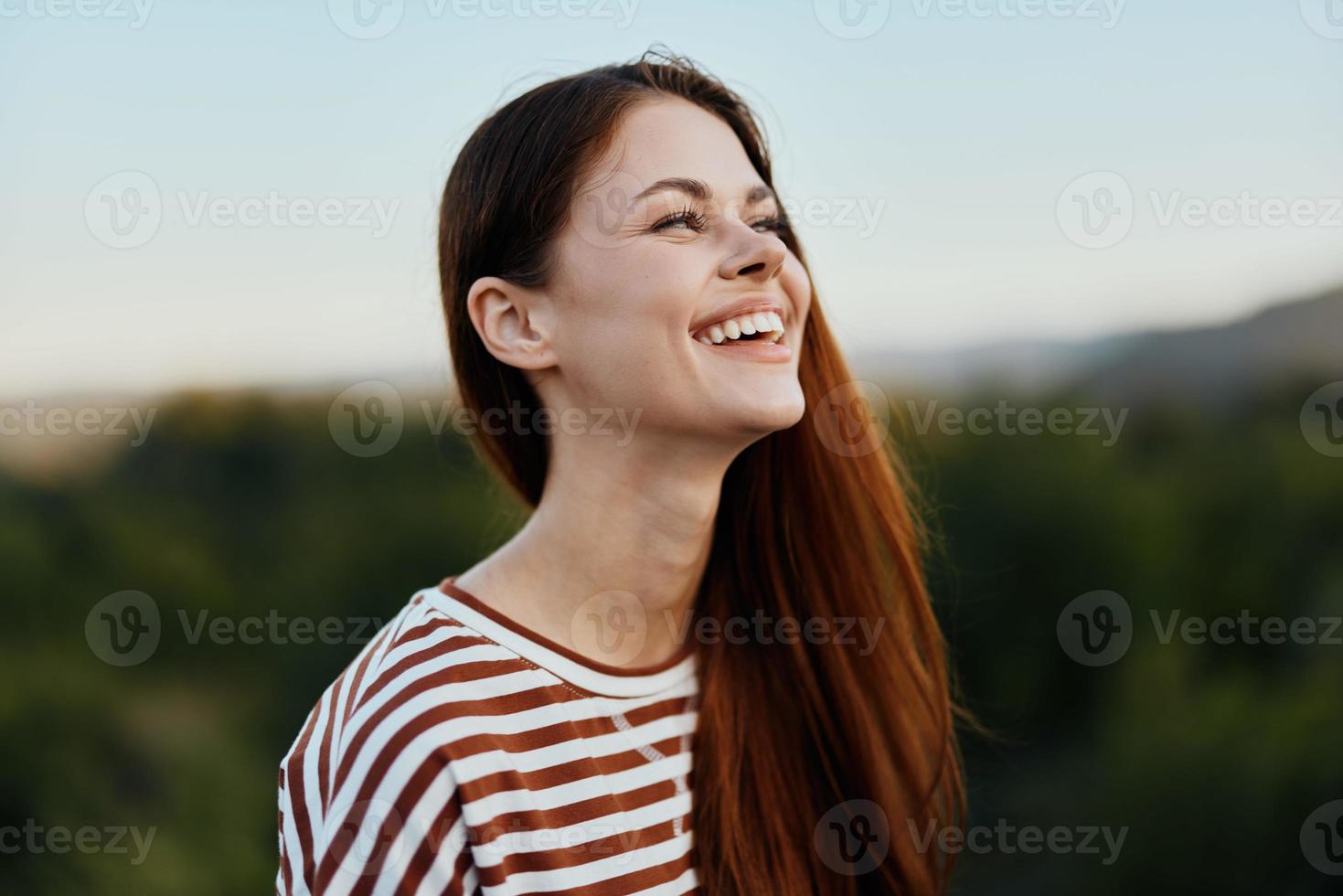 Close-up portrait of a young woman with a beautiful smile with teeth in a striped t-shirt against the background of trees photo