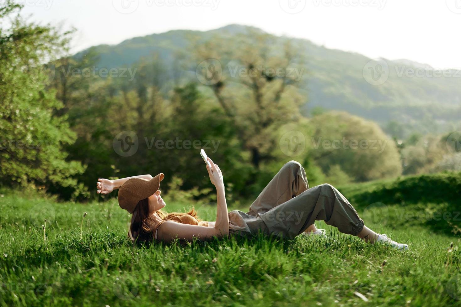 Happy woman blogger lying on the grass in the park and smiling with her phone in her hands against the backdrop of a summer natural landscape with sunlight photo