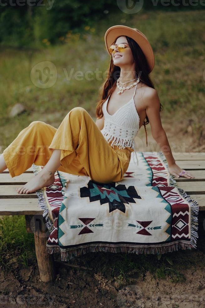 A happy girl sitting in eco clothing in nature on a plaid by the lake wearing a hat in a hippie look and smiling in wonder. Autumn Lifestyle photo