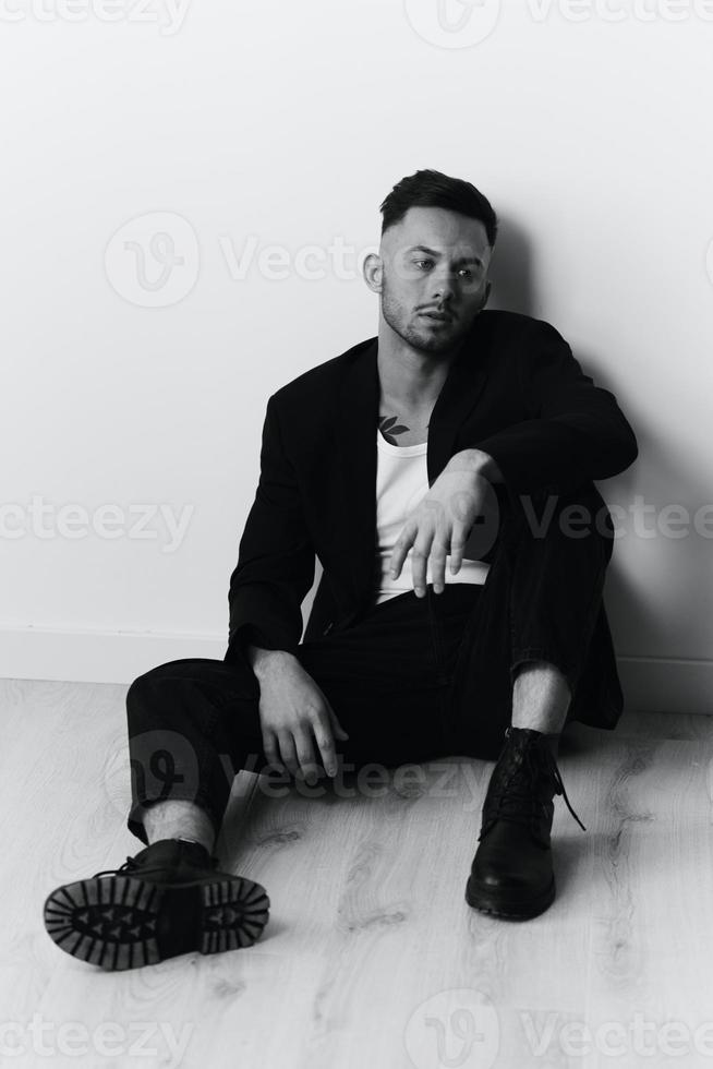 Modelling snapshots. Serious self-confident attractive handsome man in black jacket sitting on floor posing in white studio background. Black and White concept. Copy space photo