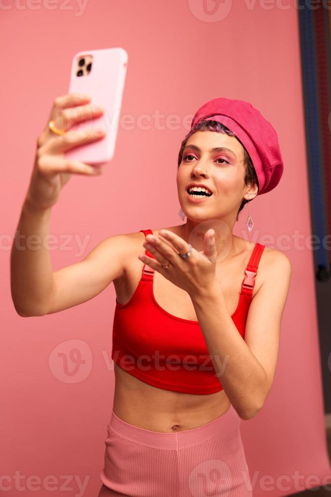 A young woman blogger with colored pink hair and a short haircut takes a picture of herself on the phone and broadcasts a smile in stylish clothes and a hat on a pink background monochrome style photo