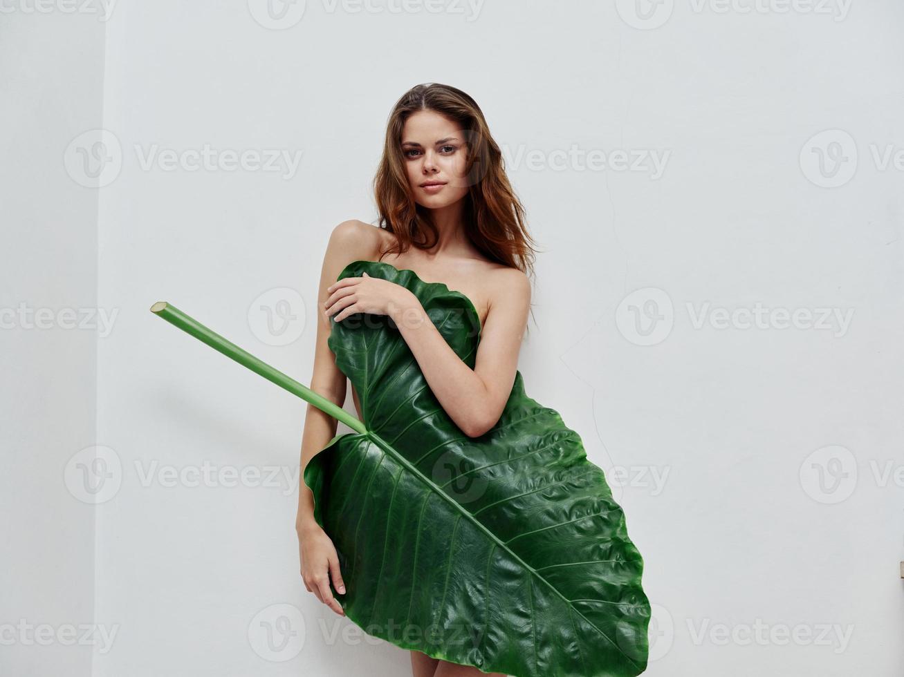 attractive woman covers naked body with green leaf light background photo