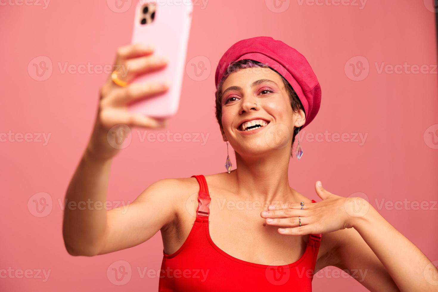 A young woman blogger with colored pink hair and a short haircut takes a picture of herself on the phone and broadcasts a smile in stylish clothes and a hat on a pink background monochrome style photo