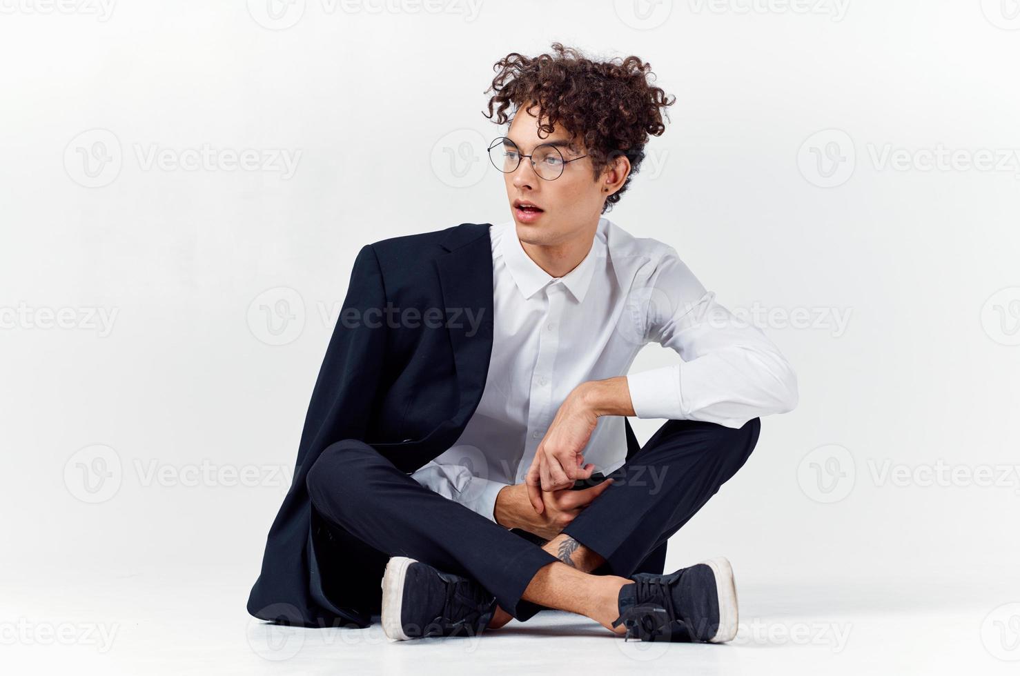 curly-haired guy in fashionable clothes and sneakers sits on the floor in a bright room in full growth photo