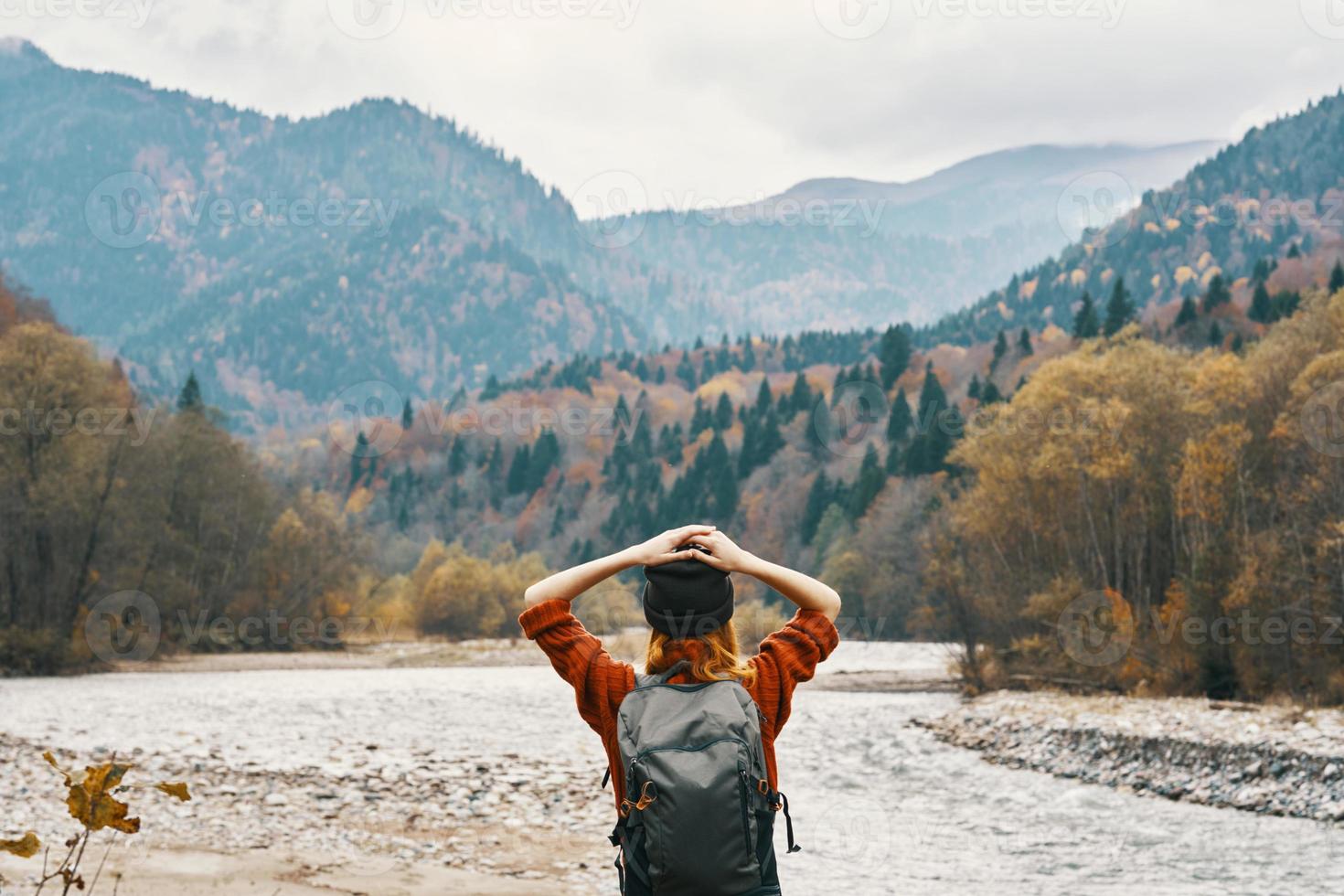 a traveler in a sweater hat with a backpack gestures with her hands and relaxes on the river bank in the mountains photo