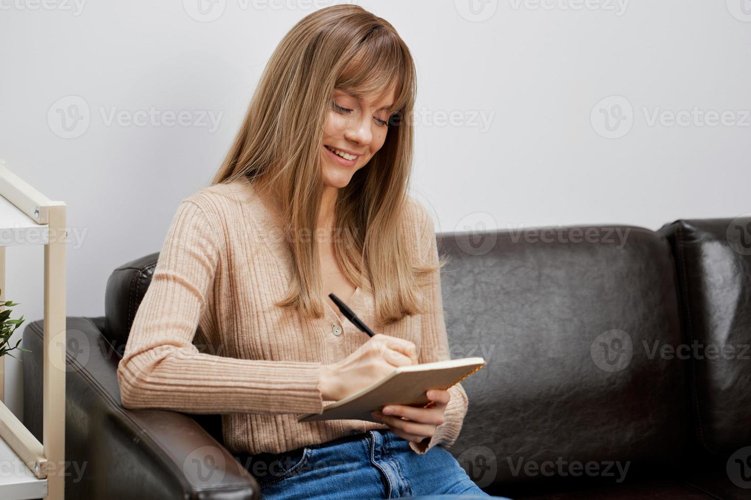 sonriente enfocado rubia estudiante dama con libro de copiar escribir letra sentado en sofá sofá a moderno vivo habitación interior. hembra cliente tener encogimiento cita en un psicológico ayuda oficina. Copiar espacio foto