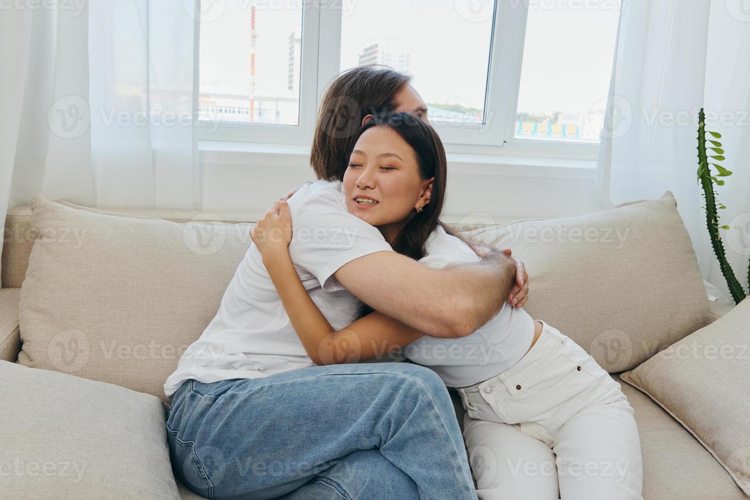 A man and a woman are sitting on the couch at home wearing white T-shirts and hugging each other with smiles. Family life lifestyles of young marrieds photo