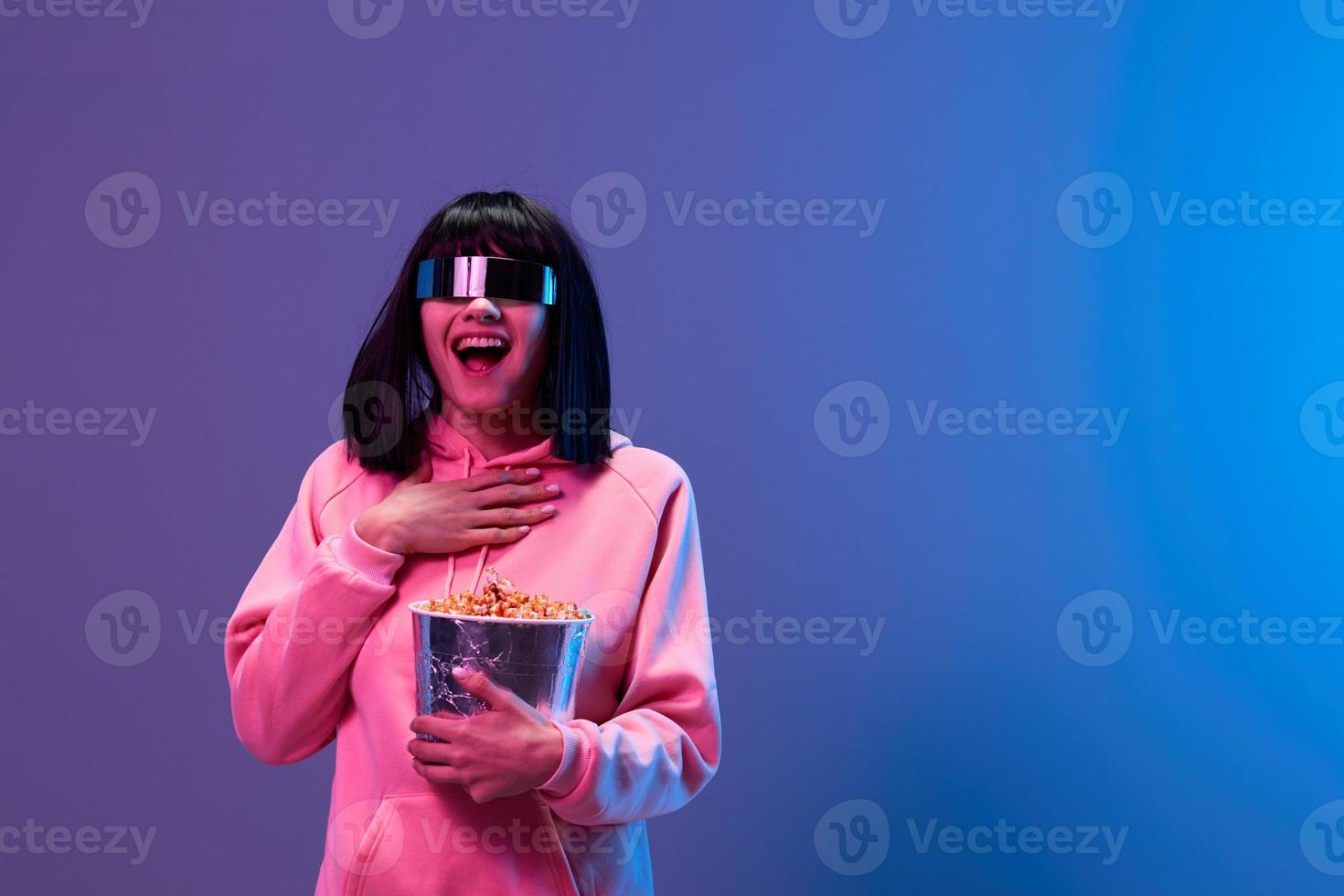 Happy excited pretty brunet woman in pink hoodie trendy specular sunglasses with popcorn hold hand on chest posing isolated in blue violet color light background. Neon party Cinema concept. Copy space photo