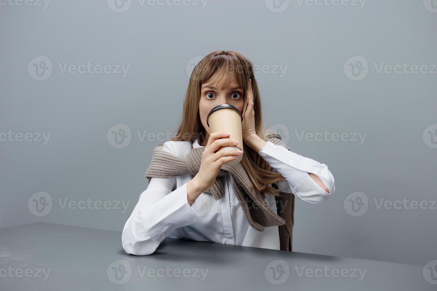 Excited young blonde student lady freelancer in warm sweater drink takeaway coffee look at camera sitting at workplace in gray modern home office. Coffee break Lover Concept. Copy space. Cool offer photo