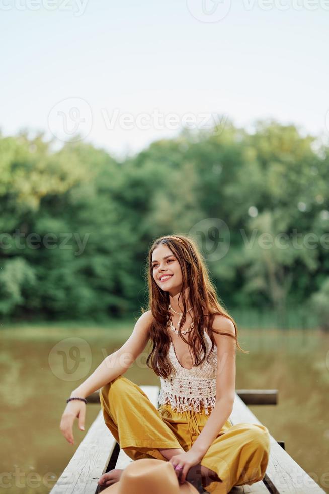 un hippie mujer se sienta en un puente por un lago en un naturaleza viaje y sonrisas en ropa ecológica. el concepto de eco-activista y naturaleza conservación foto