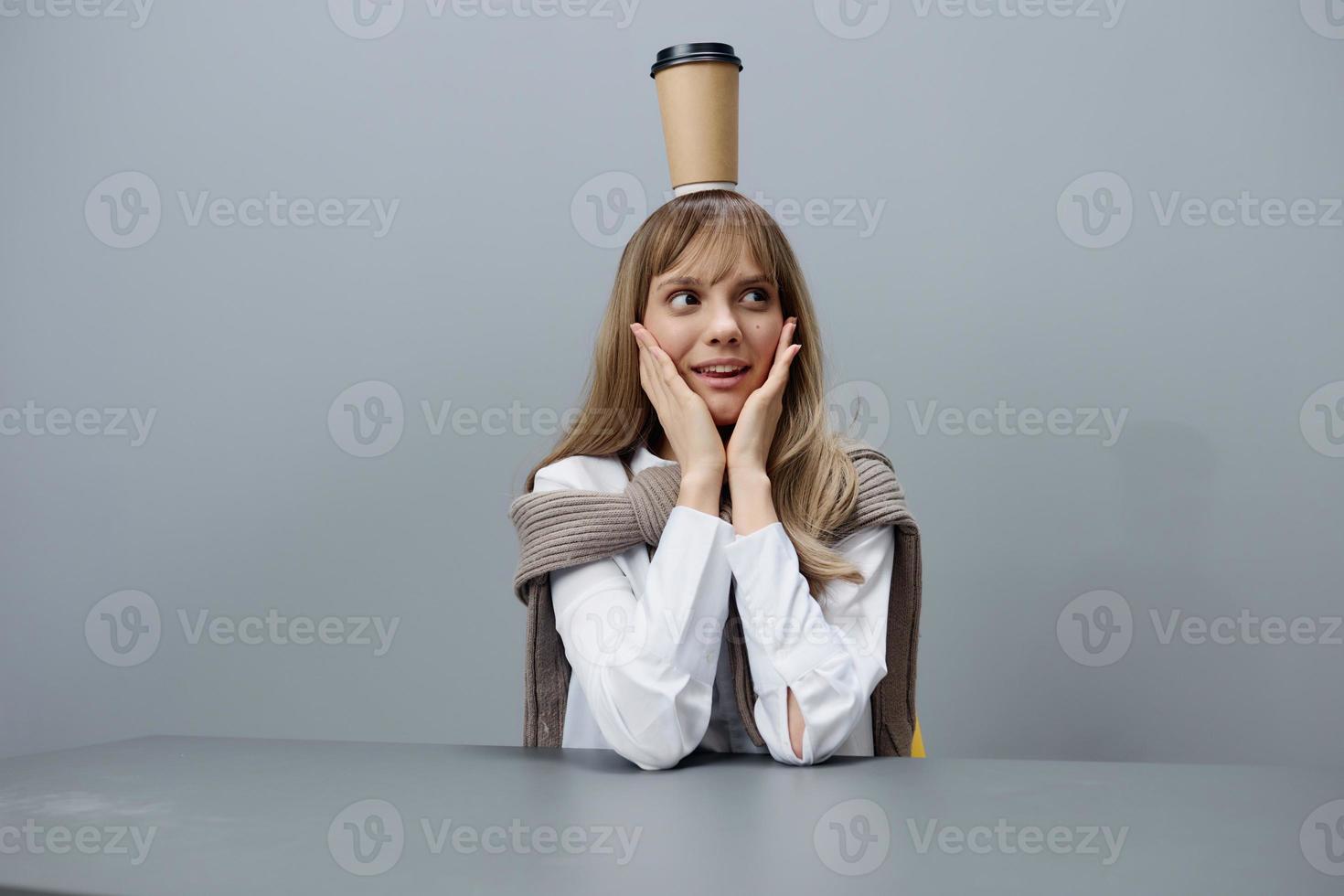 Funny young blonde student lady freelancer in warm sweater with takeaway coffee on head touch cheeks sitting at workplace in gray modern home office. Coffee break Lover Concept. Copy space. Cool offer photo