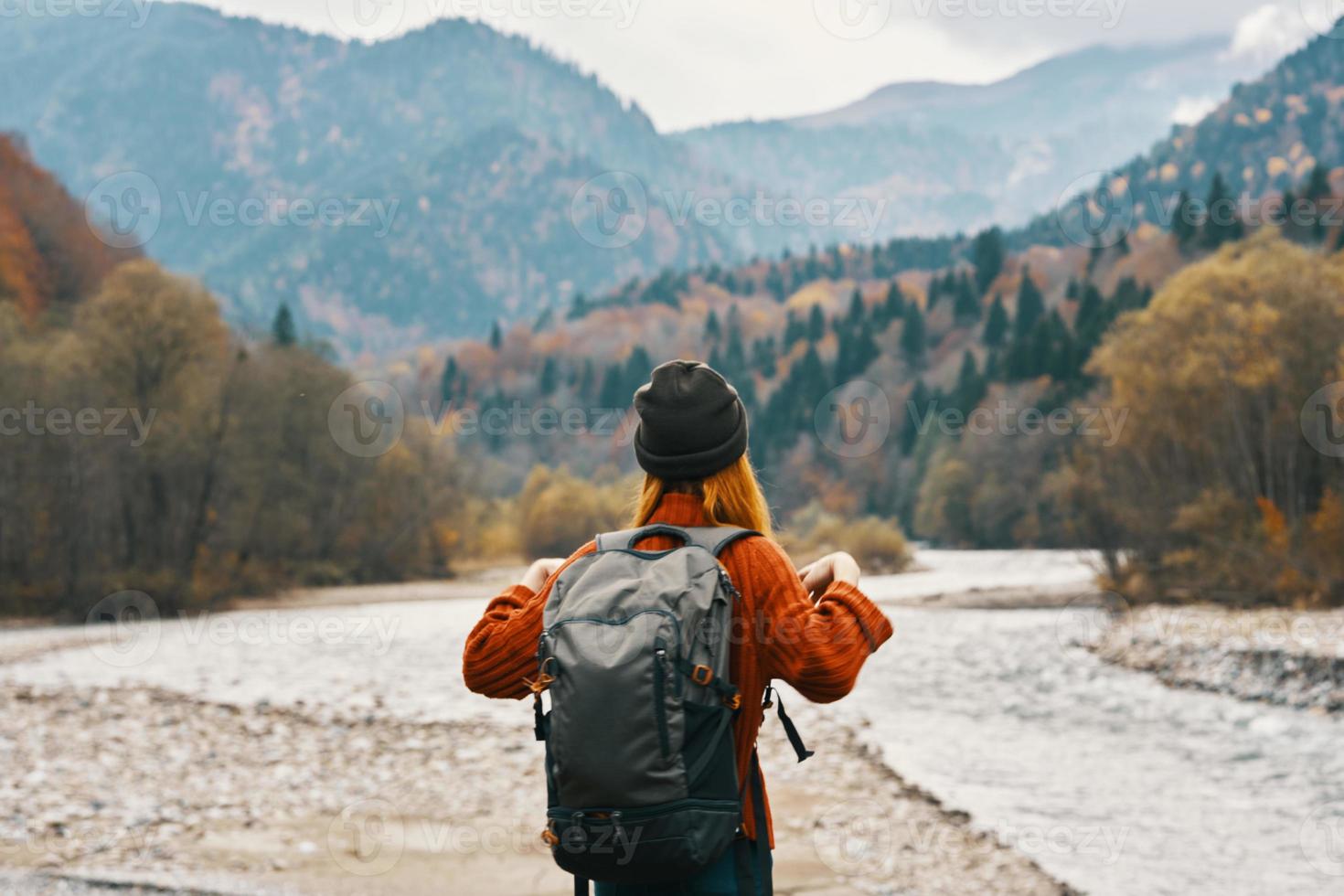 joyful woman tourist with a backpack looks at the mountains near the river on the bank photo