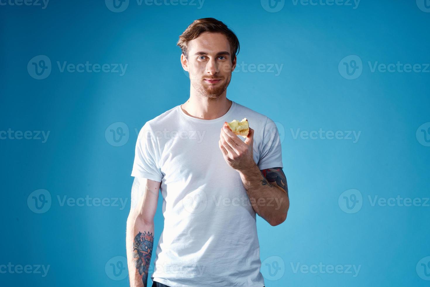 cute guy with a tattoo and in a white t-shirt on a blue background eating apples cropped view photo