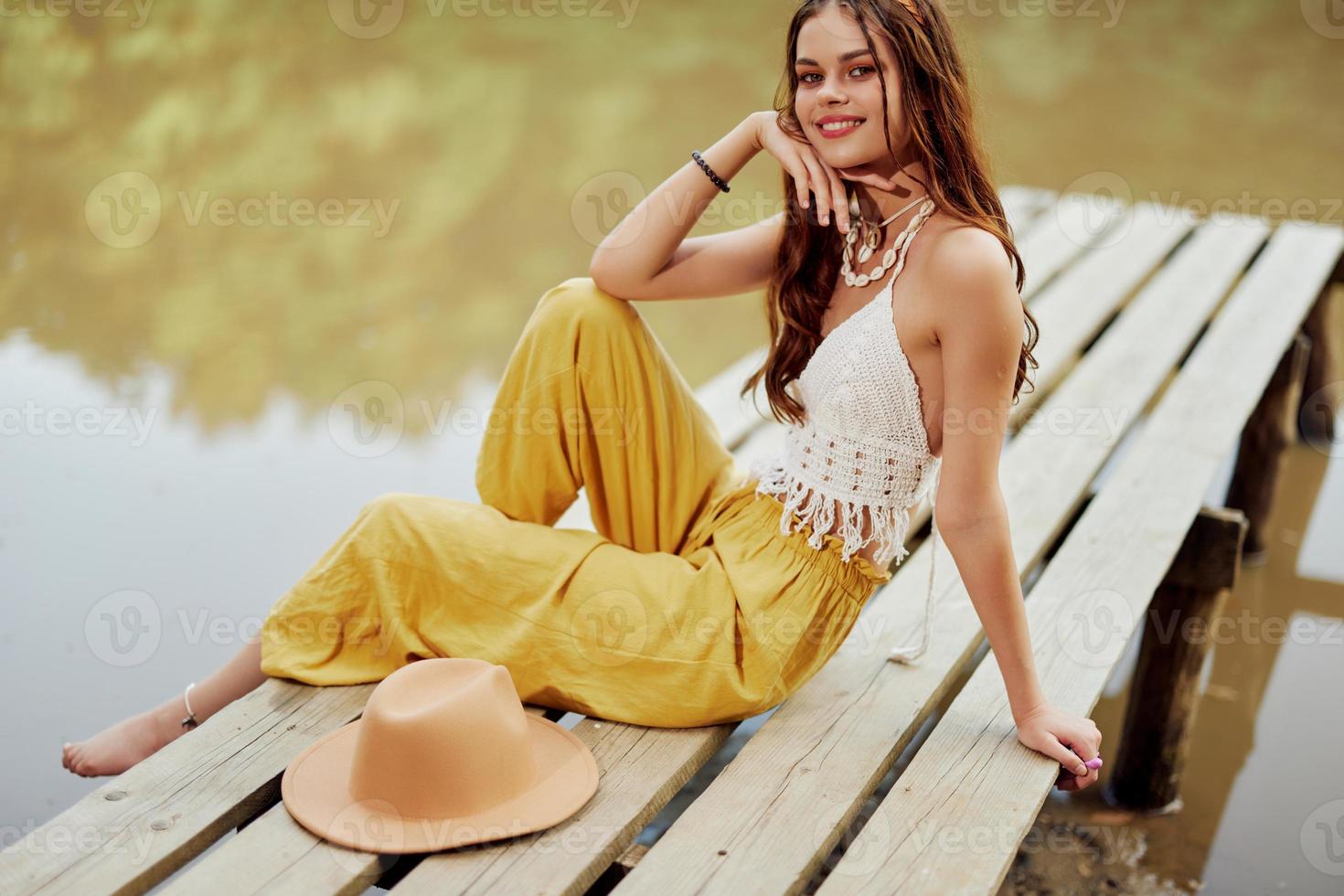 A young hippie woman sits on a lake bridge wearing stylish eco clothes and smiling photo