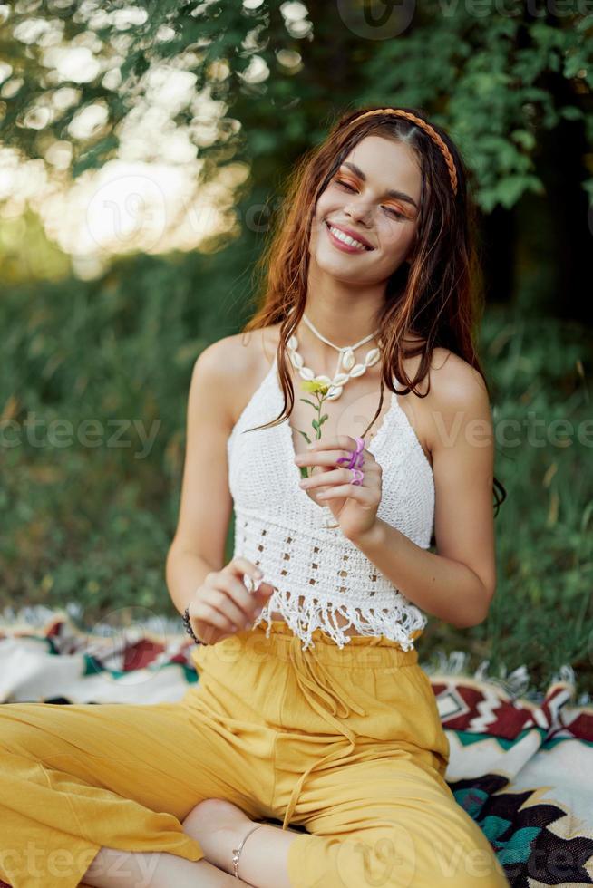 Young hippie woman sitting in nature on a plaid with a flower in her hands and looking at the camera smiling sincerely in eco-clothing in autumn in the sunset photo