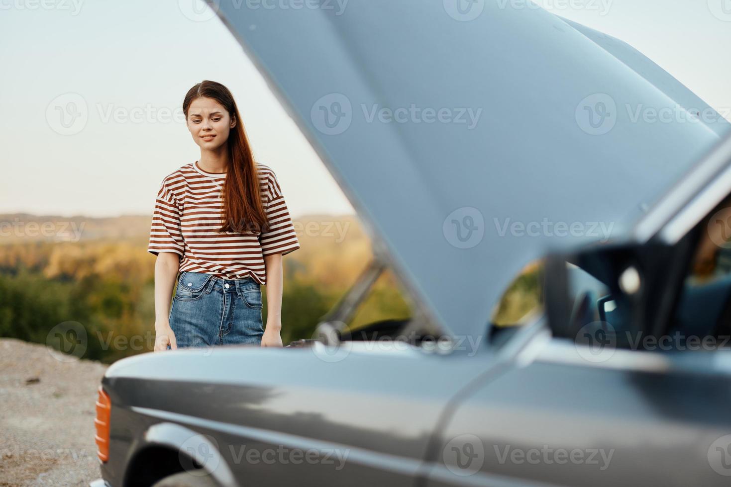 Woman traveler standing near the car with the hood open and looking for the cause of the car breakdown alone without men on the road in the countryside photo