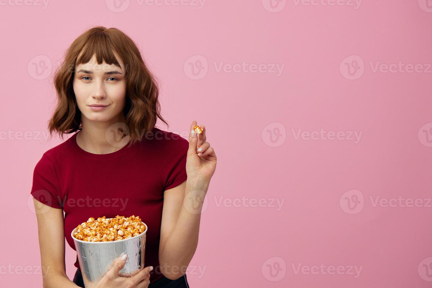 Smiling cheerful pretty cute redhead lady in red shirt with popcorn ready to watch movie posing isolated on over pink studio background. Copy space Banner. Fashion Cinema concept. Entertainment offer photo