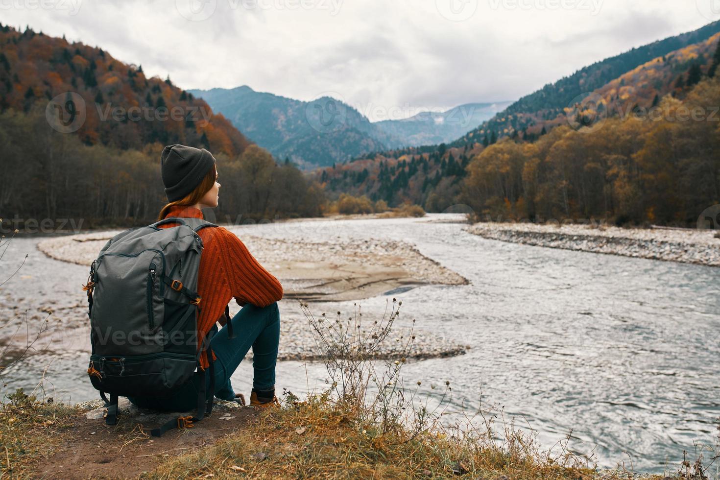 woman with a backpack in a jacket and a hat on the river bank in the mountains side view photo