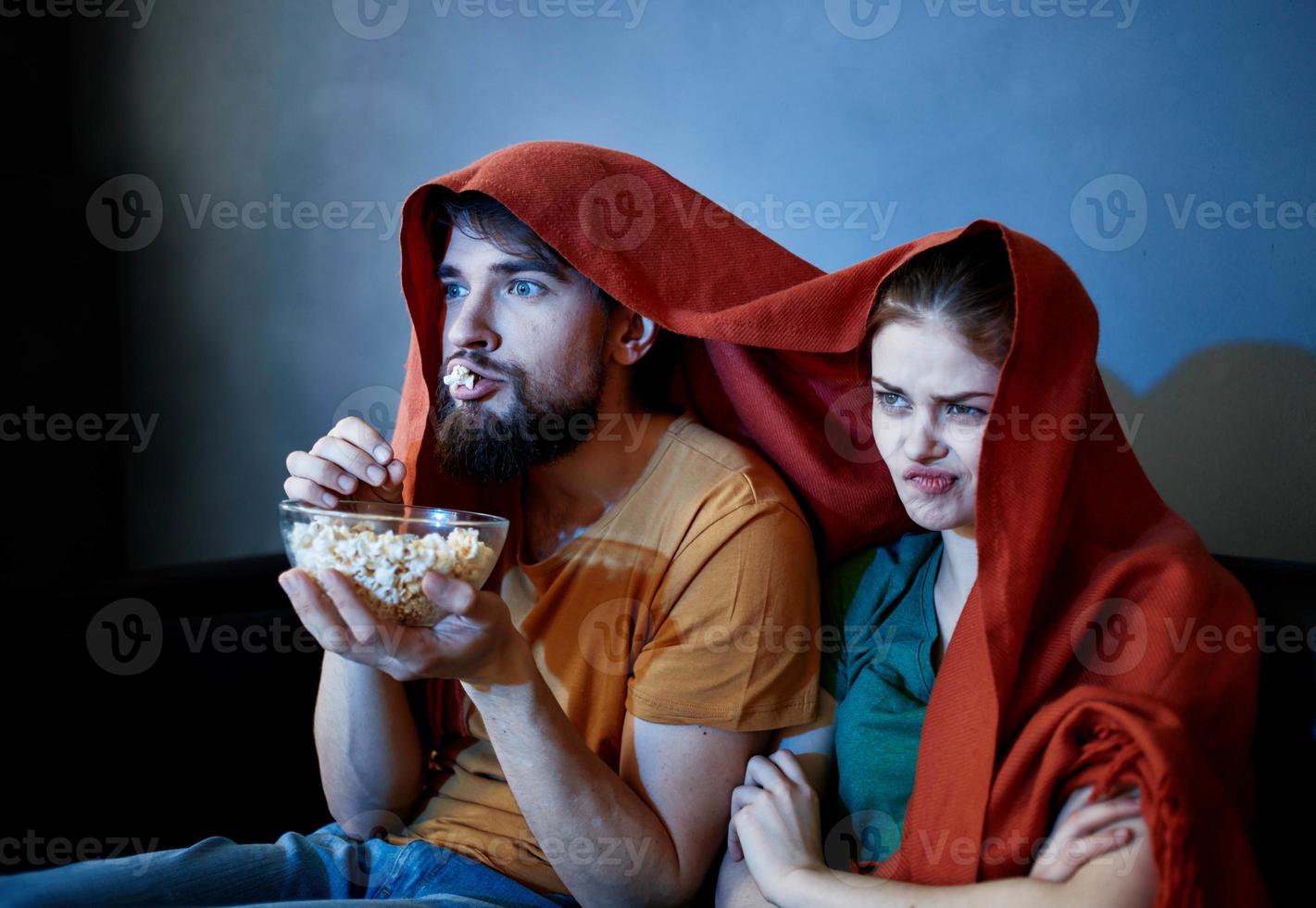 A man with a plate of popcorn and an emotional woman under a red blanket on the couch photo