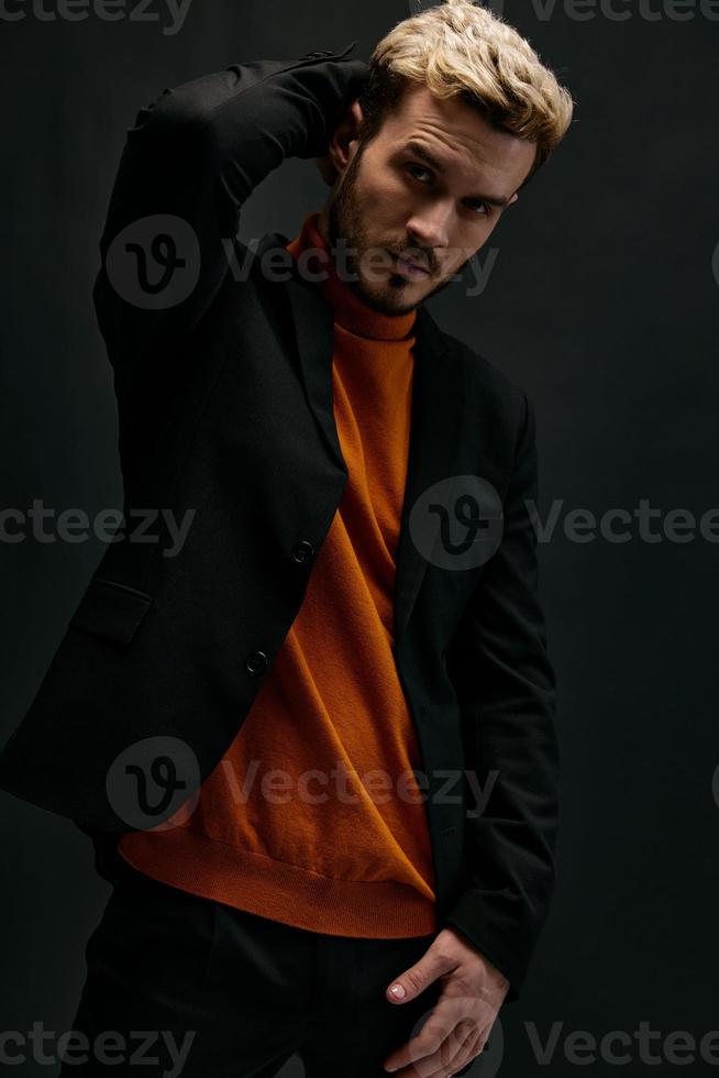 elegante hombre modelo en un suéter y Saco sostiene su mano detrás su cabeza en un oscuro antecedentes Copiar espacio foto