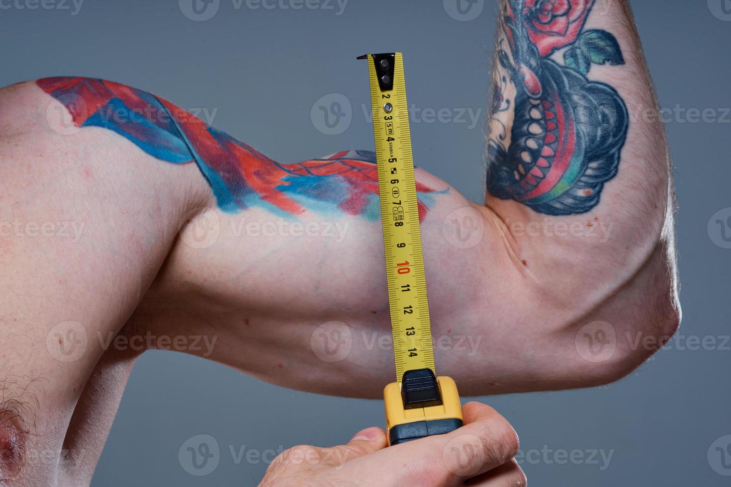 guy measures arm muscles with centimeters on a gray background and a multicolored tattoo bodybuilder fitness photo
