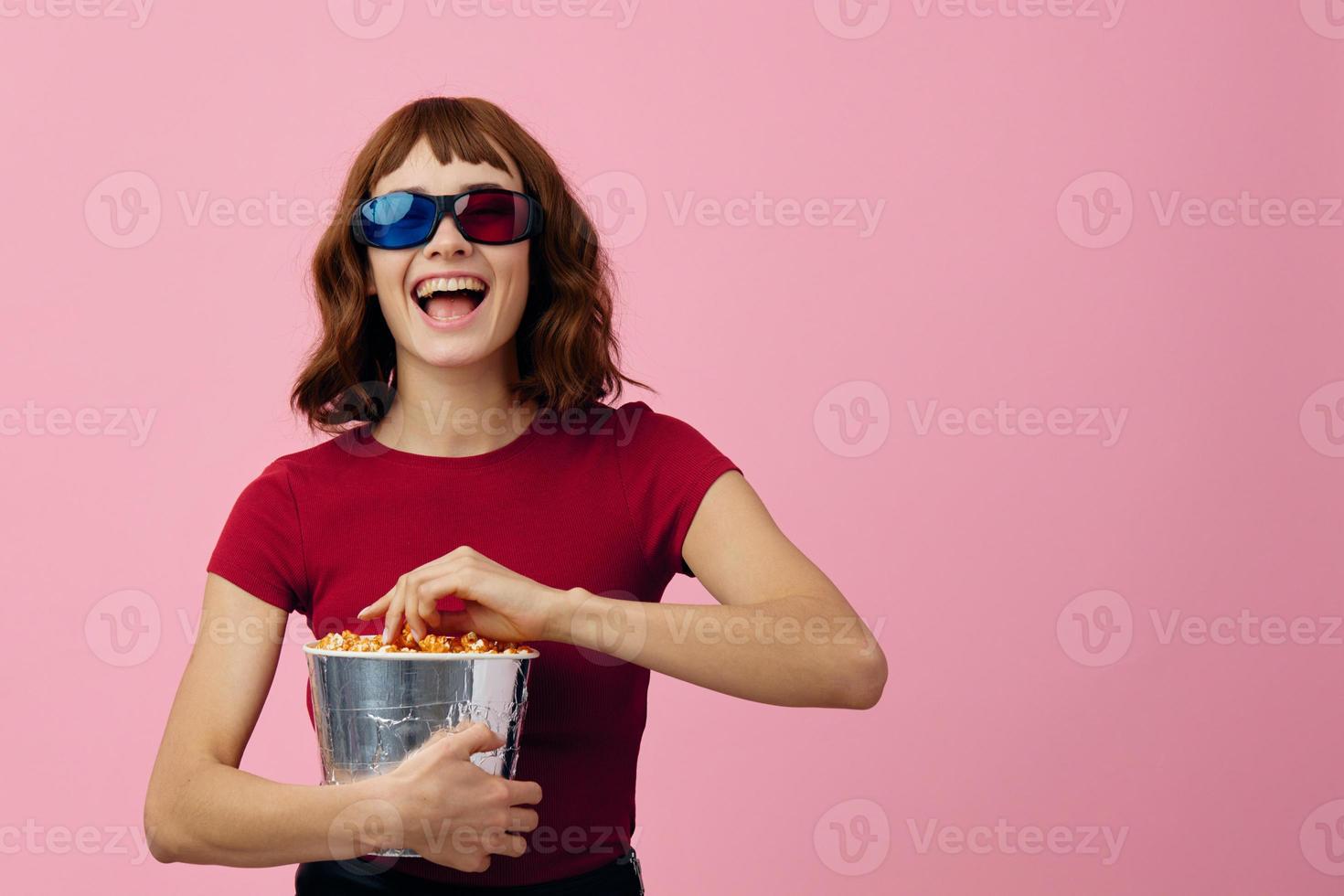 lleno de alegría contento linda pelirrojo dama en rojo camiseta 3d lentes con palomitas de maiz reloj película posando aislado en terminado rosado estudio antecedentes. Copiar espacio bandera. Moda cine concepto. entretenimiento oferta foto