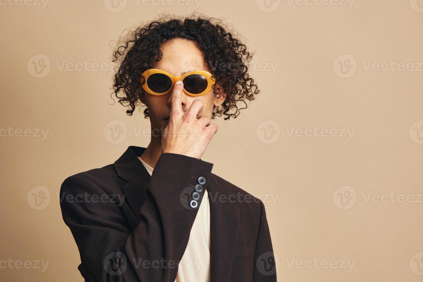 Demonstrative awesome tanned curly man in classic brown jacket trendy sunglasses posing isolated on over beige pastel background. Fashion New Collection offer. Retro style concept. Free place for adDemonstrative awesome tanned curly man in classic brown j photo