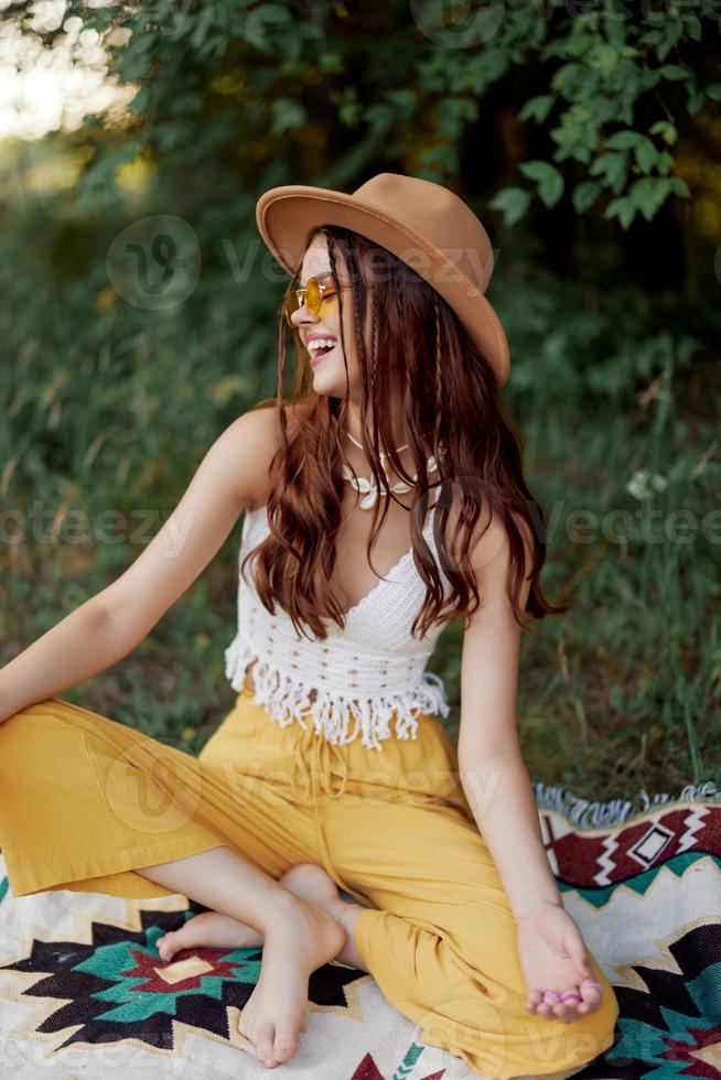 A young hippie woman in eco-clothing has fun laughing and cringing sitting on a plaid outside in the fall with a hat and yellow sunglasses. Travel lifestyle photo