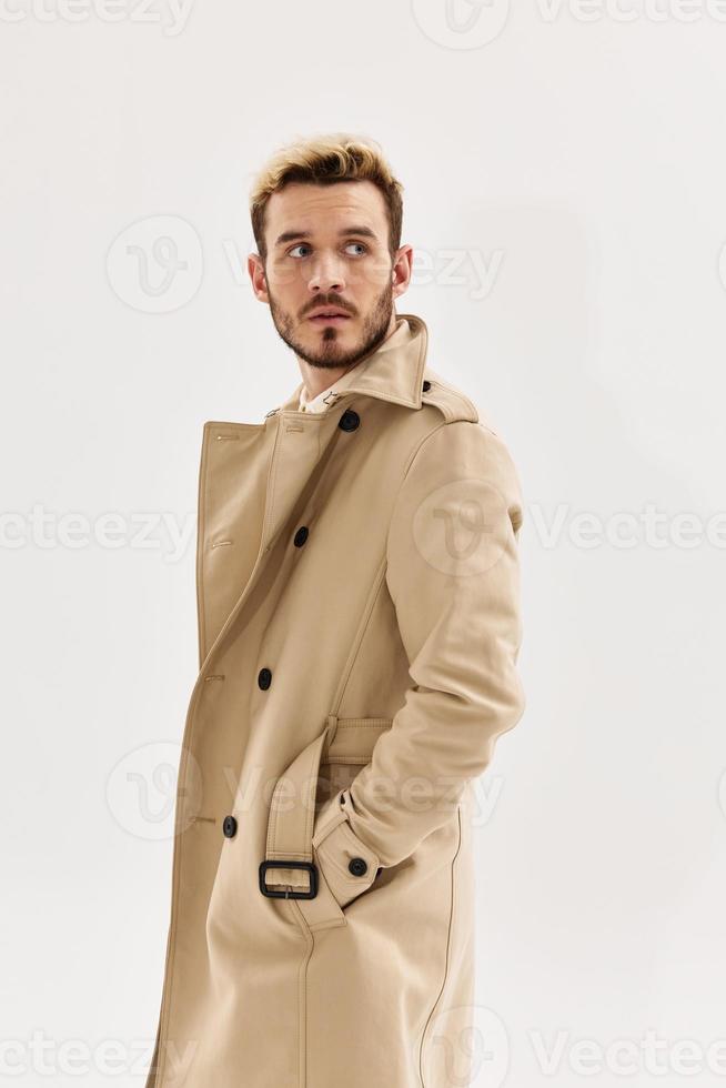 a man in a beige coat holds his hands in his pocket a look to the side light background photo